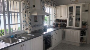 Cuina amb armaris blancs, un vàter d'acer inoxidable, forn encastrat i finestra amb cortines florals.