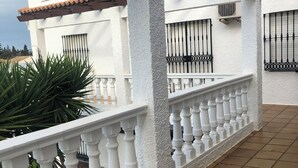 Una zona exterior coberta amb una barana blanca, un paviment de rajoles i un edifici amb teulada i finestres reixades.