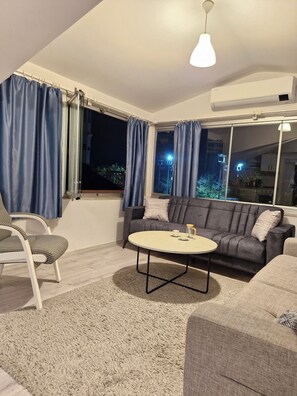 A living room with a sofa, a round coffee table, a chair, and a large window with curtains. - Gorgeous apartment close to the sea with private beach (Antalya)