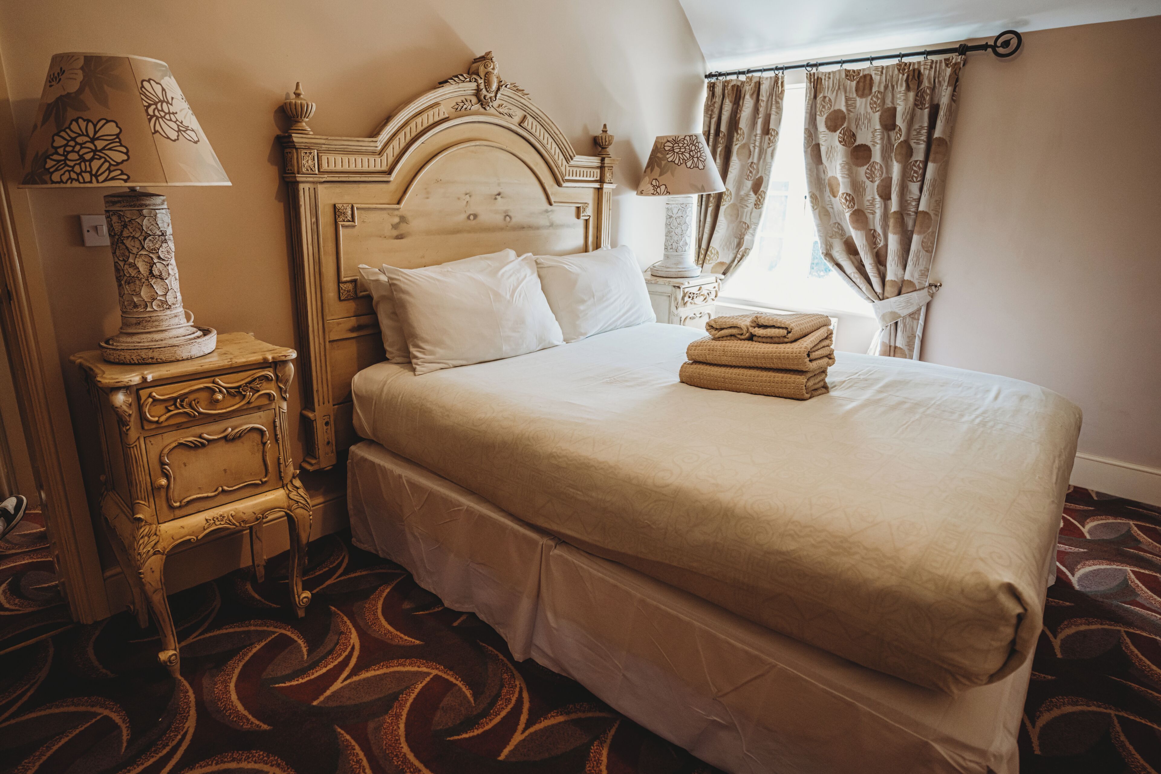 A hotel room with a large bed, a nightstand with a lamp, a headboard, and a window with curtains.
