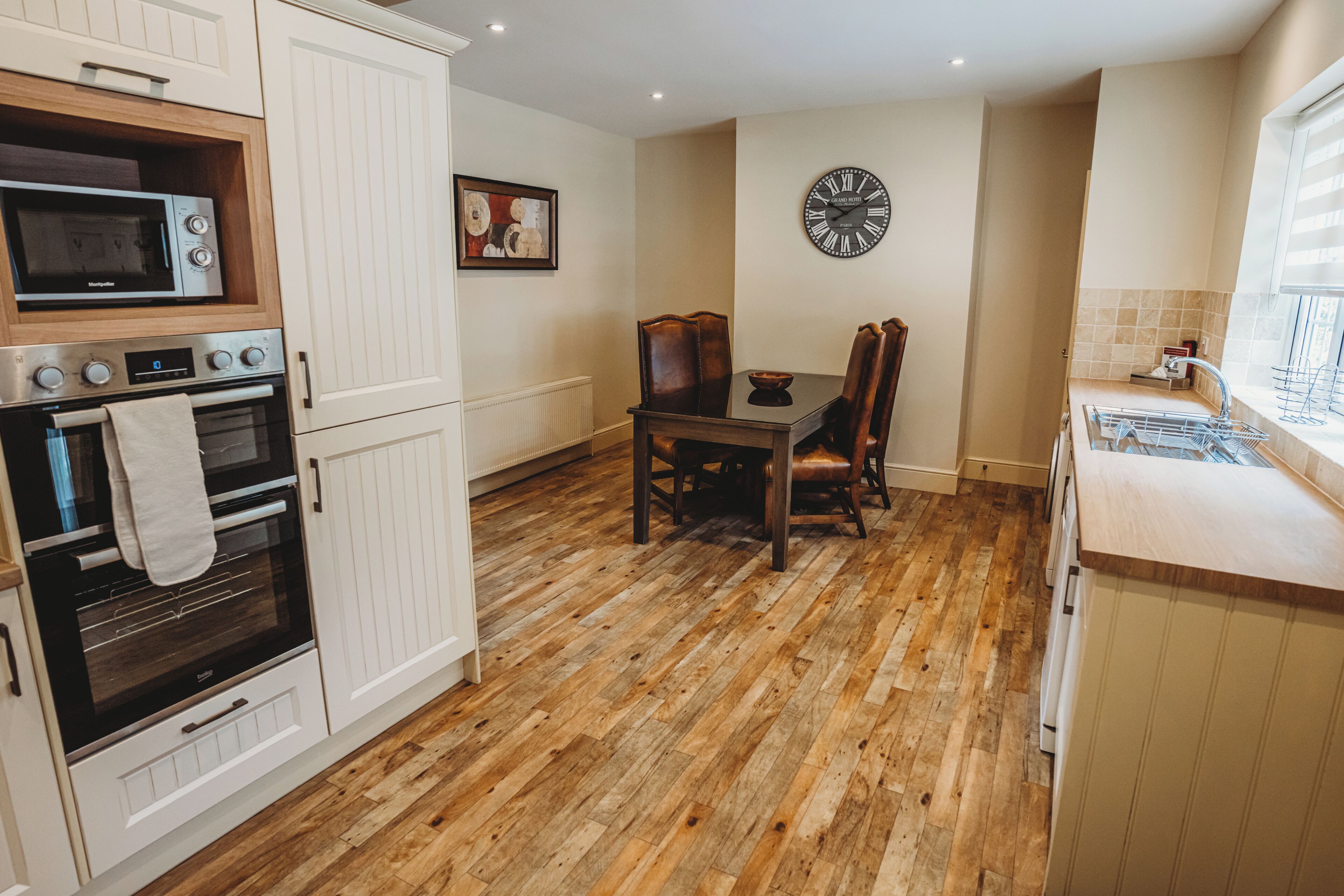 Una cocina con muebles blancos, piso de madera, una mesa de comedor con sillas, un reloj de pared y una ventana. Se observa una toalla colgada en la puerta del horno.
