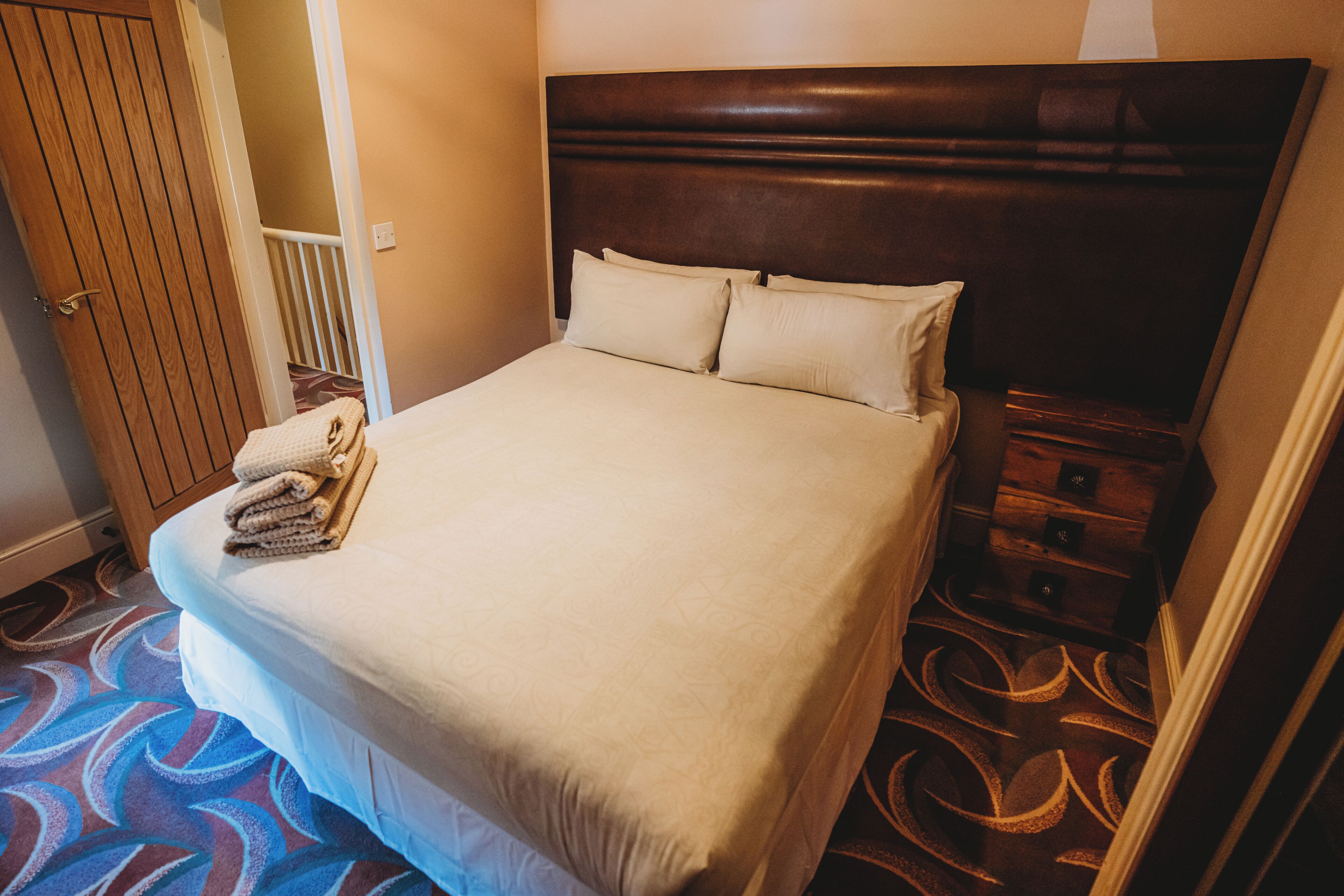 Una cama bien hecha con cabecera, una pila de toallas dobladas y una mesita de noche de madera con cajones.