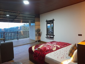 A bedroom with a large bed, a standing fan, a mirror, and a balcony with a view of the outdoors.