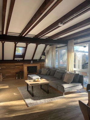 A modern living room with a sloped ceiling, a fireplace, a sofa, and a view of the outdoors. - Chiemgau Getaway: Vacation Home with Mountain Views (Siegsdorf)
