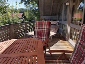 Eine Holzdiele mit Tisch und Stühlen, eine Glastür und Blick auf Bäume und Gebäude.