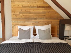 Ein Schlafzimmer mit einem Bettgestell aus Holzleisten, einem Bett mit weißer Bettwäsche und zwei Kissen, einem dunkel strukturierten Teppich und einer Lampe auf einem Nachttisch.