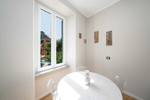 Ein Zimmer mit rundem Tisch, Stühlen, einem Fenster mit Blick auf Gebäude und Grünflächen sowie Wandbildern.
