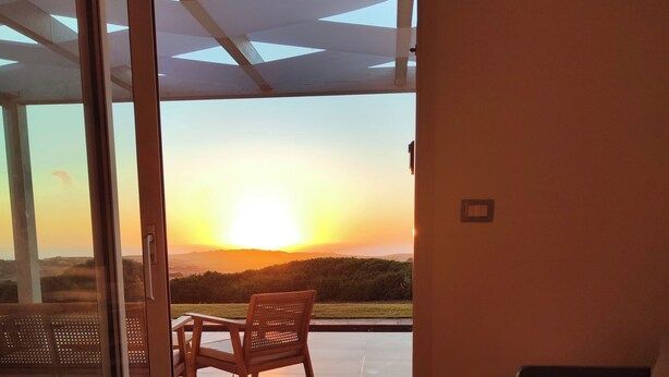 A room with a sliding glass door leading to an outdoor patio with a view of the sunset over a hilly landscape.