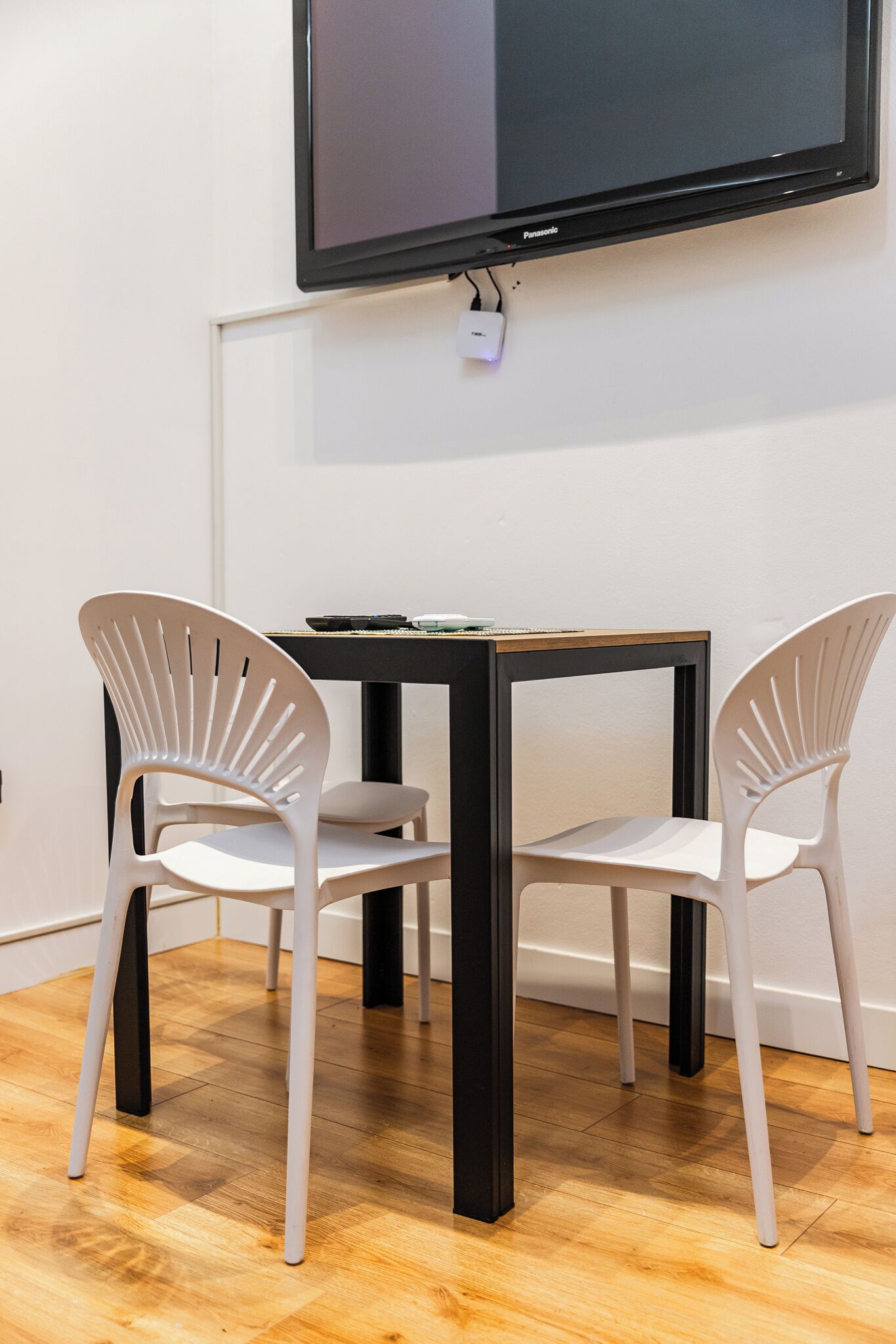 A room with a wooden floor, a black table, two white chairs, and a flat-screen TV mounted on the wall.
