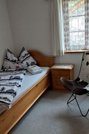 A bedroom with a single bed, a nightstand, a chair, and a window with lace curtains. - Ferienwohnungen Teuriblick A&B, (Splügen), by Interhome (Splügen)