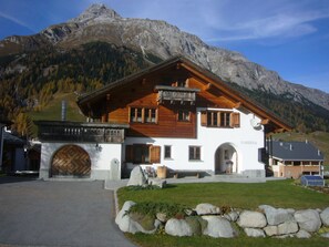 A chalet-style building with a wooden facade, large windows, and a prominent entrance with a wooden door. - Ferienwohnungen Teuriblick A&B, (Splügen), by Interhome (Splügen)