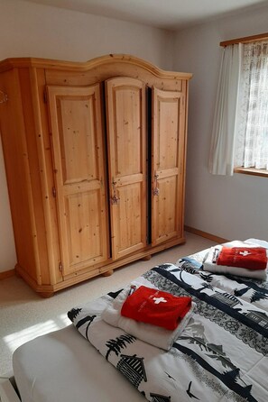 A bedroom with a wooden wardrobe, a bed with patterned bedding, and a window with a sheer curtain. - Ferienwohnungen Teuriblick A&B, (Splügen), by Interhome (Splügen)