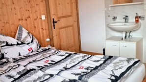 Slaapkamer met een houten deur, een bed met beddengoed met een patroon, een witte nachtkastje, en een badkamer met een wastafel en spiegel.
