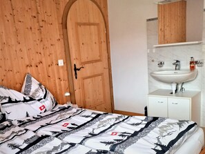 A bedroom with a wooden door, a bed with patterned bedding, a white bedside table, and a bathroom with a sink and mirror. - Ferienwohnungen Teuriblick A&B, (Splügen), by Interhome (Splügen)