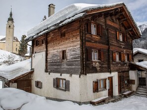 A traditional wooden building with a snow-covered roof, a visible chimney, and a bell tower in the background. - Ferienwohnungen Felder, (Splügen), by Interhome (Splügen)