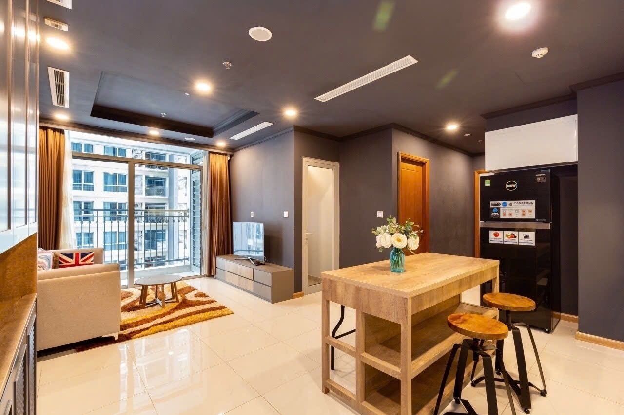 A modern living room with a sofa, a coffee table, and a TV. There is a kitchen area with a fridge, a wooden table, and two stools.