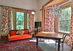 Una habitación con una mesa de madera, un sofá, un televisor y ventanas grandes con cortinas florales.