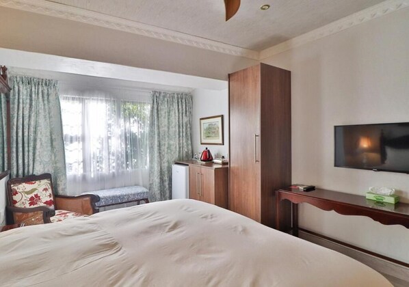 A hotel room with a large bed, a television, a desk, a chair, a wardrobe, and a window with curtains. - Victoria House (Ficksburg)