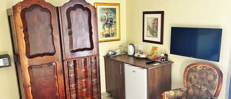 A room with a wooden cabinet, a white refrigerator, a floral armchair, a television, and a painting on the wall.