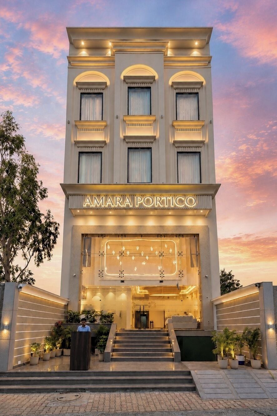 Un bâtiment moderne sur lequel on peut lire l’inscription « AMARA PORTICO ». Il se distingue par une entrée imposante, avec des marches menant à une porte, le tout agrémenté de plantes en pot.
