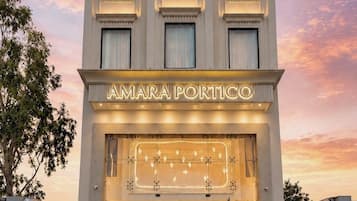 Un bâtiment moderne sur lequel on peut lire l’inscription « AMARA PORTICO ». Il se distingue par une entrée imposante, avec des marches menant à une porte, le tout agrémenté de plantes en pot.