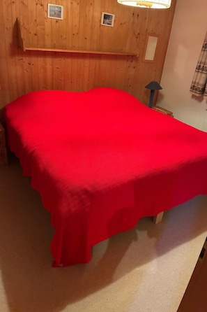 A room with a bed covered in a red blanket, wooden walls, a small shelf, and a lamp.