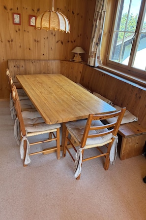 A wooden dining table with cushioned chairs, a hanging light fixture, and a window with curtains.