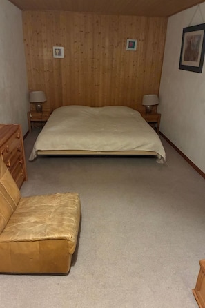 Quarto com cama, cabeceira de madeira, duas luminárias de apoio, cômoda de madeira e sofá marrom.