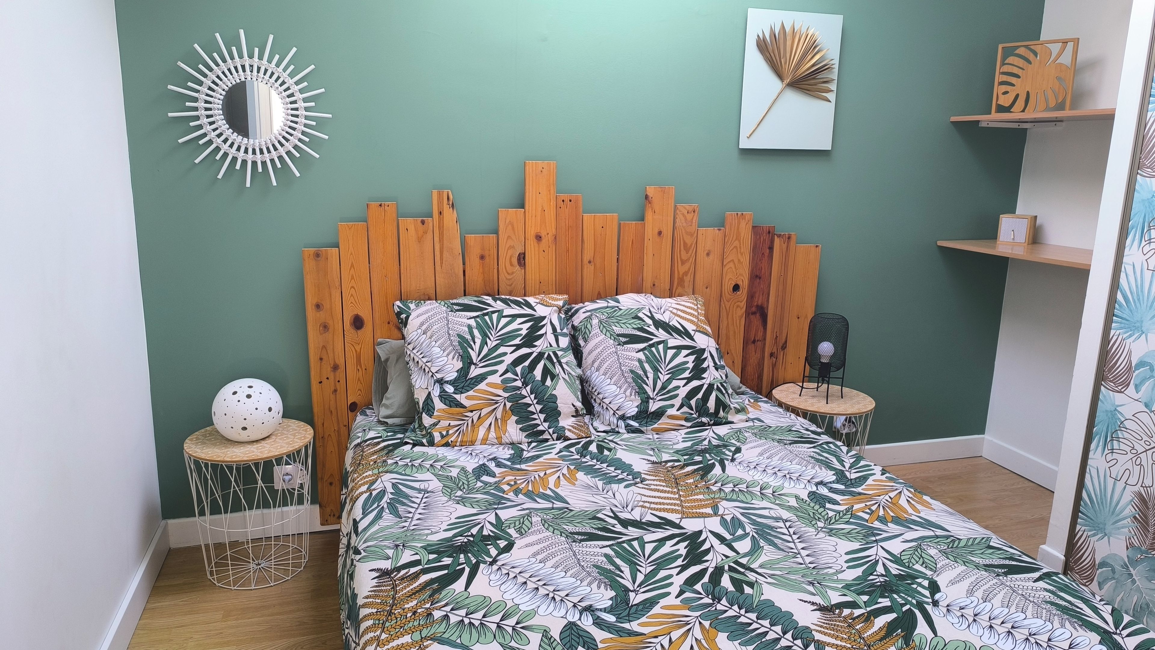A bedroom with a wooden headboard, a bed with a leaf-patterned bedspread, a round side table, a decorative mirror, and a wall-mounted shelf with a plant.