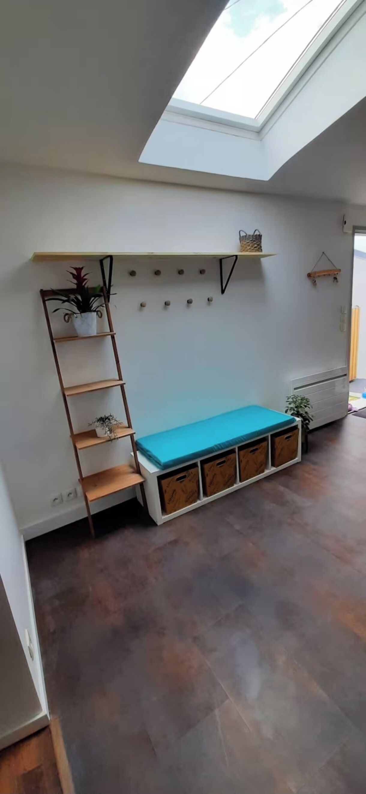 Ein Zimmer mit Oberlicht, einer blauen Matratze, einer Holzleiter und einem an der Wand befestigten Regal mit einer Pflanze.