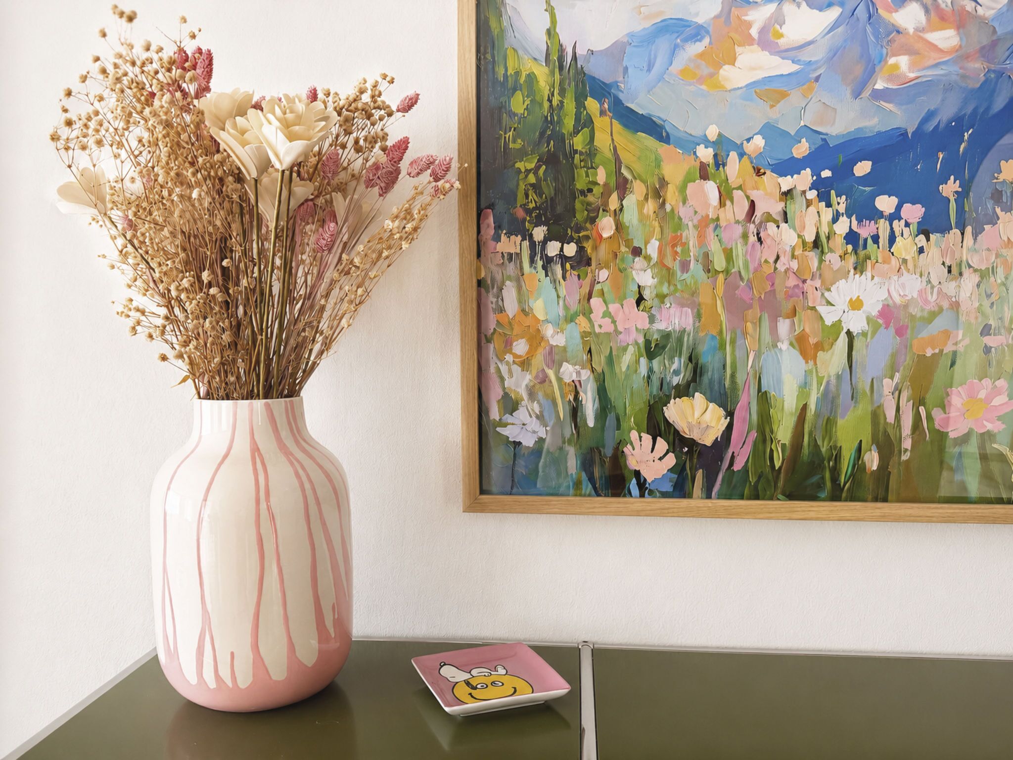 Un jarrón rosa con flores secas sobre una mesa, junto a un cuadro enmarcado.