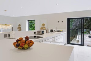A modern kitchen with a central island, a bowl of fruit, and a view of the outdoors through large sliding doors. - Perched above Matlock Bath, 37 Steps is a stylish upside-down home designed to make the most of its incredible views across the valley. With a sociable open-plan living space, four ensuite bedrooms, and a showstopping terrace with hot tub. (Matlock Bath)