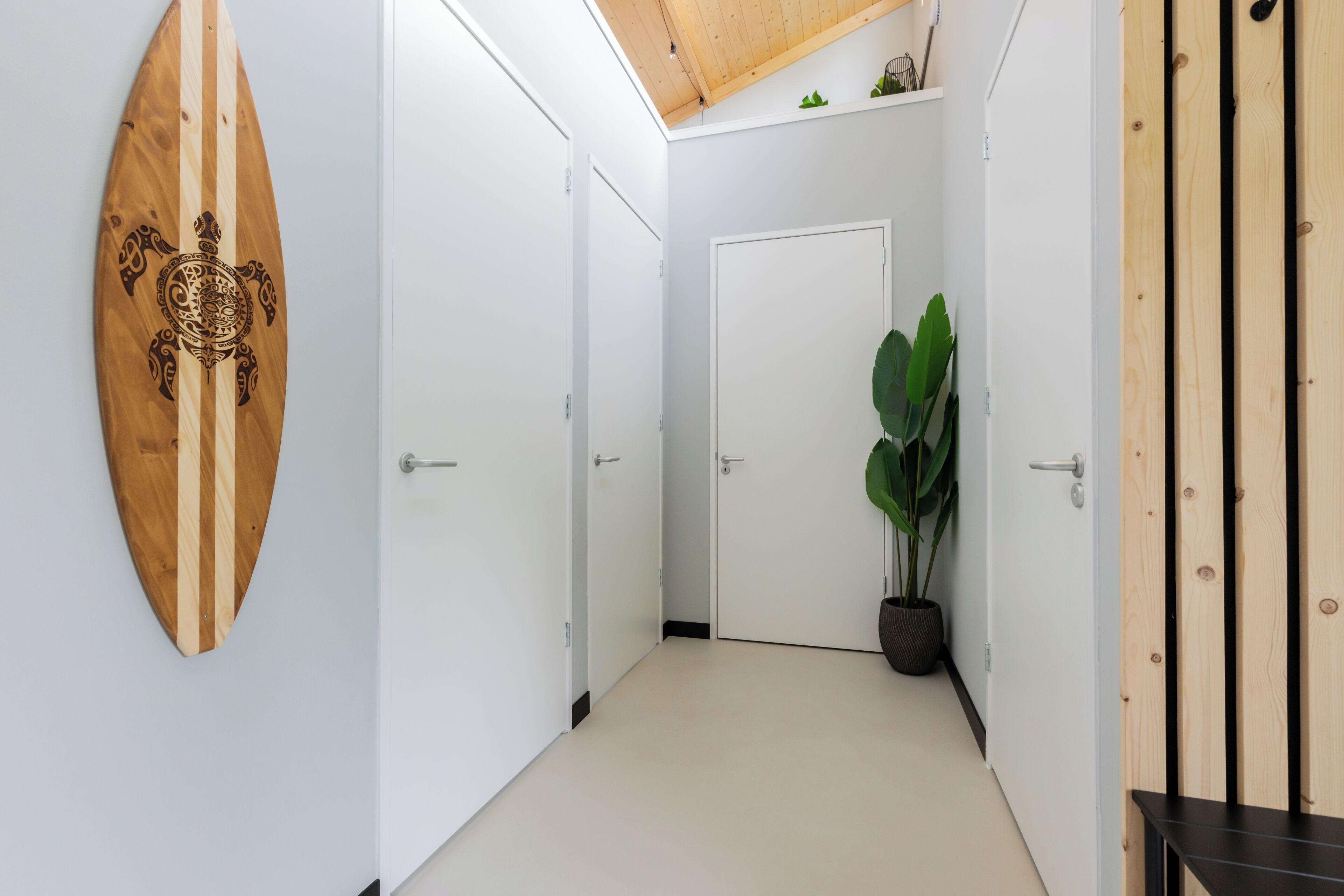 A hallway with white walls, a wooden surfboard decoration, a potted plant, and a door at the end.