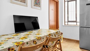 Un comedor con una mesa cubierta con mantel floral, dos sillas, una puerta de madera, una ventana con vistas y un refrigerador.