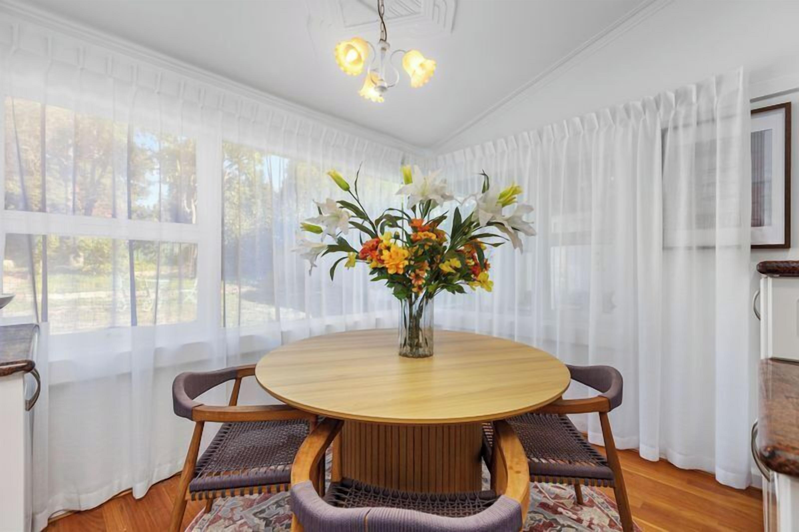 A dining area with a round table, chairs, a vase of flowers, and a view through sheer curtains.