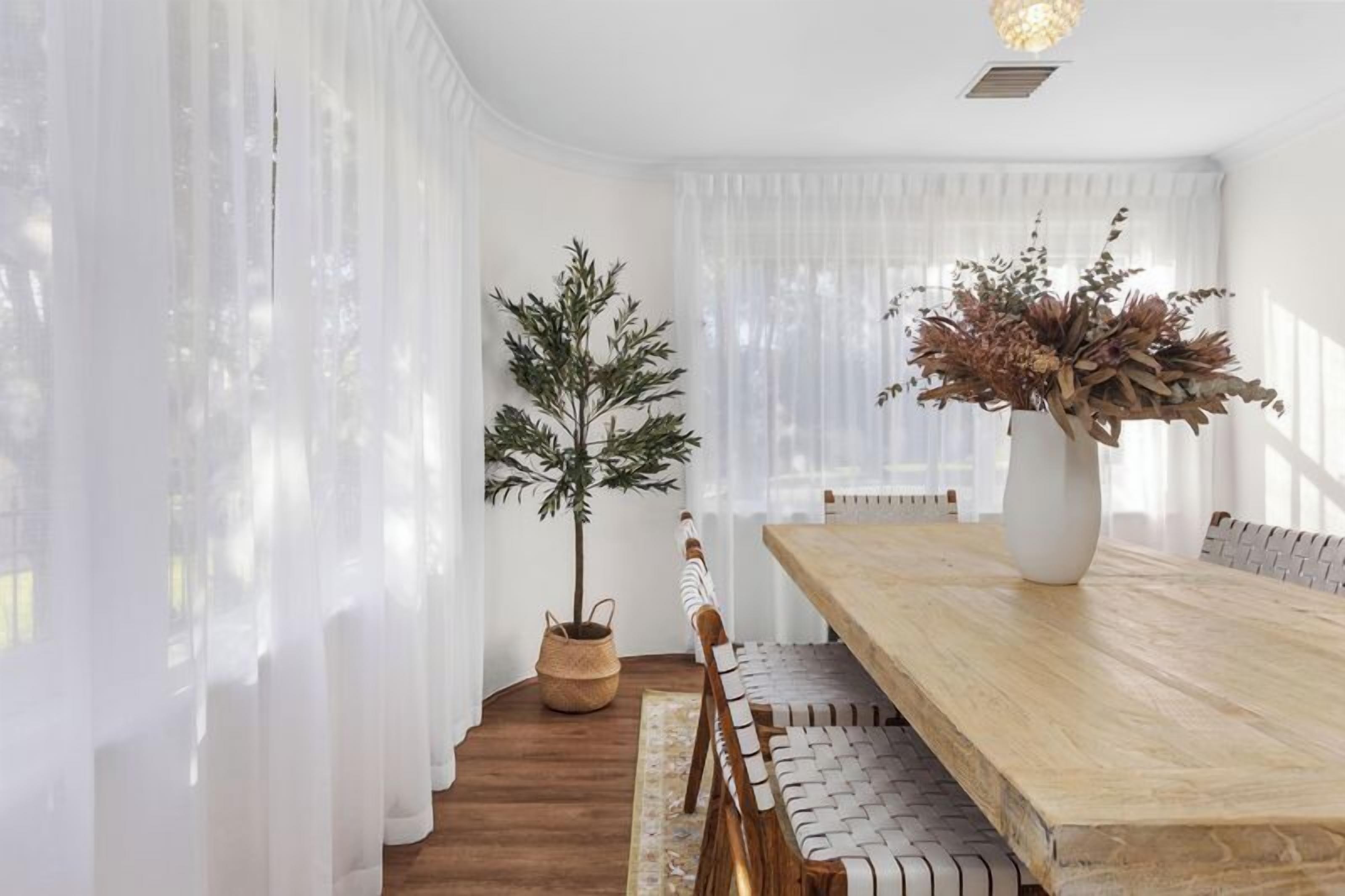 A dining area with a wooden table, white curtains, a potted plant, and a vase with dried flowers.