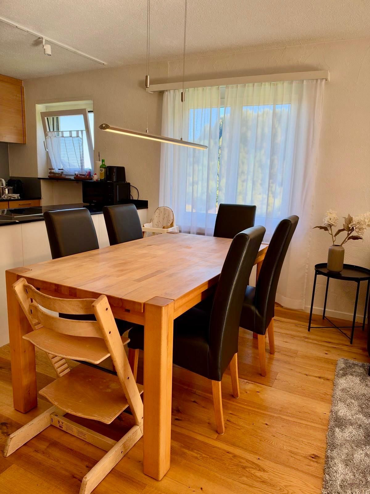 A dining area with a wooden table and chairs, a kitchen in the background, and a window with curtains.