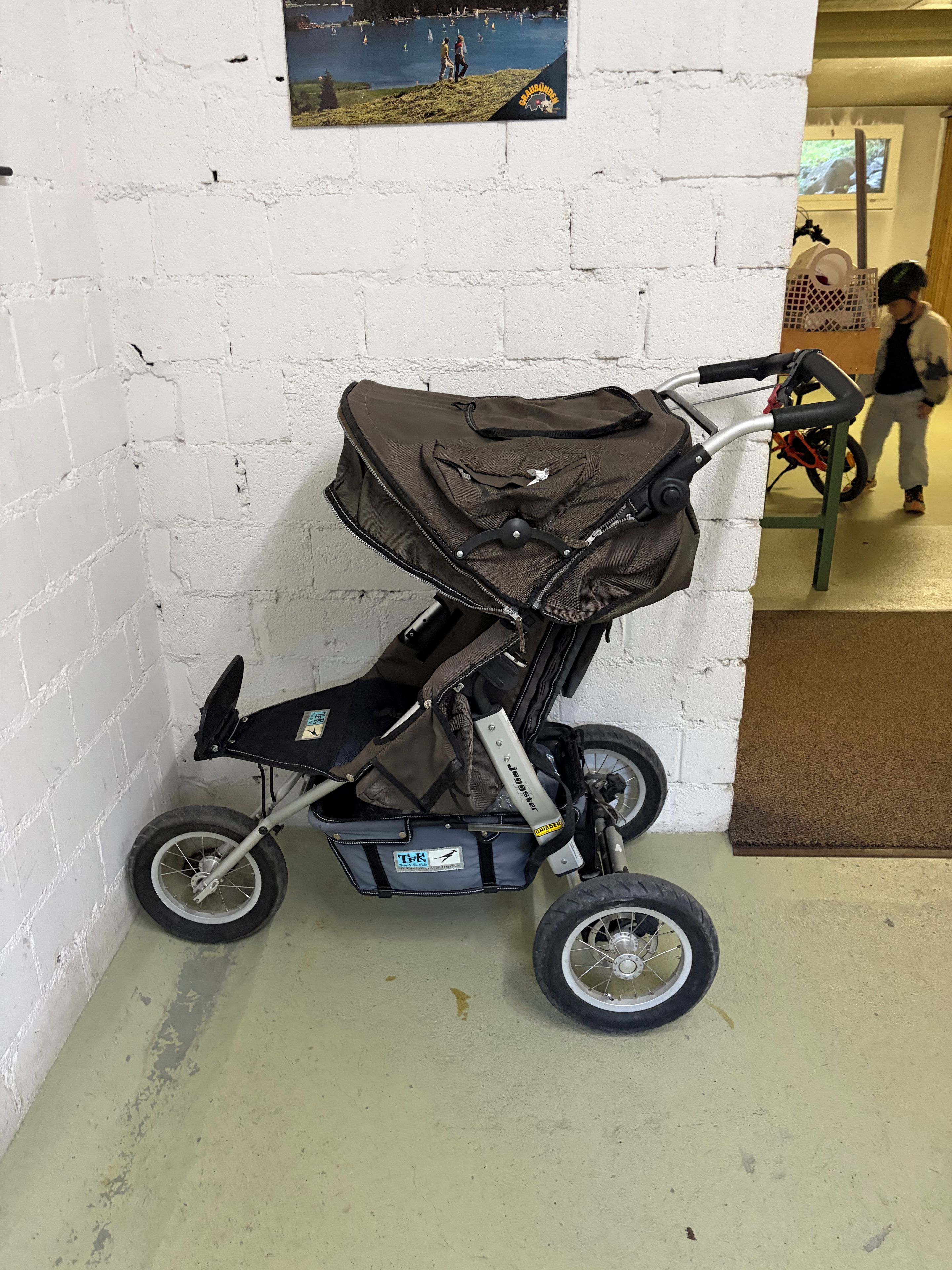 A stroller with a black seat and canopy, attached to a bicycle, against a white brick wall.