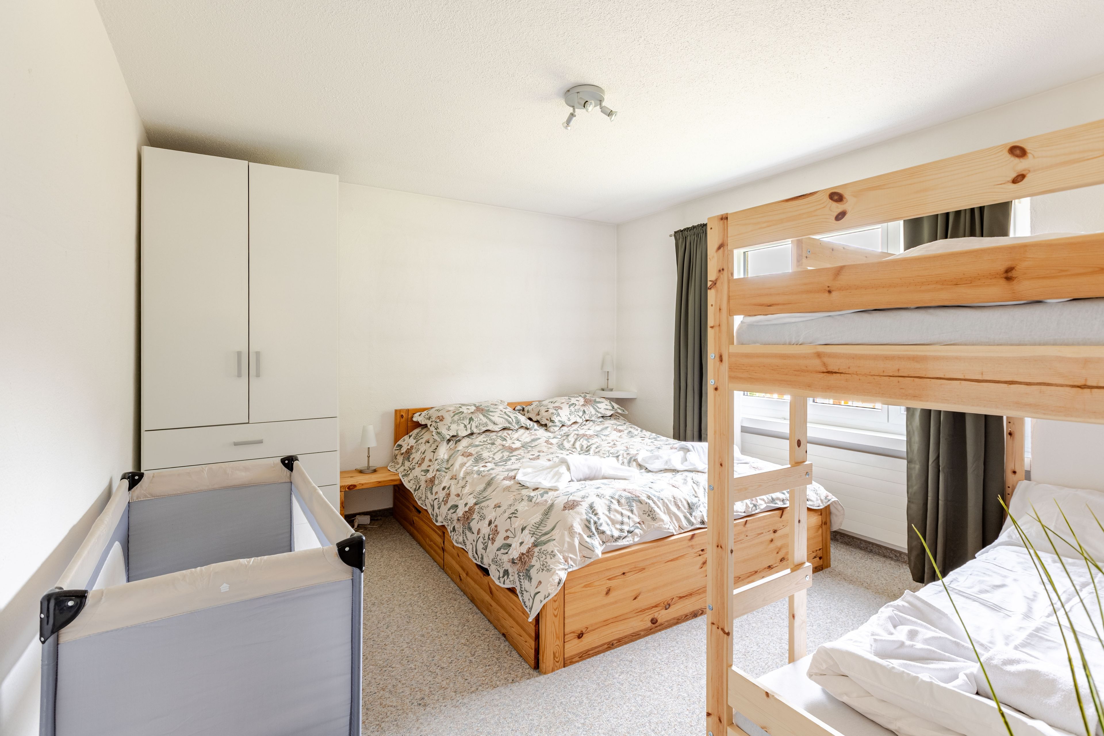 A bunk bed with a wooden frame, a single bed with a floral bedspread, a white wardrobe, and a small bedside table with a lamp.