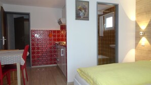 Pequeña habitación con cama, una mesa y una silla. Cuenta con un baño con ducha e inodoro. La pared es de baldosas rojas y tiene una ventana.