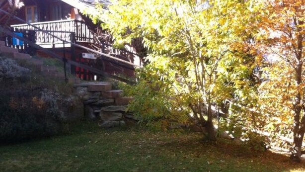 A stone staircase leading to a wooden deck with a railing, surrounded by grass and trees with autumn leaves. - Alicia, (Grächen), 1901 by Interhome (Grächen)