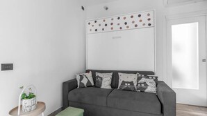 Ett modernt vardagsrum med en grå soffa, mönstrade kuddar, ett litet sidobord med en krukväxt och en vit vägg med ett färgglatt prickmönster.