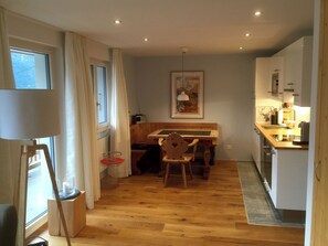 Eine kompakte Küche mit Holzboden, einem Esstisch mit Stühlen, einem kleinen Kühlschrank und einem Fenster mit Vorhängen.