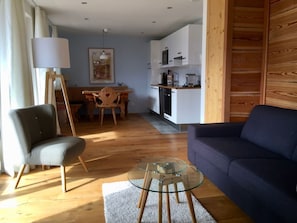 Un salon avec un canapé, une table basse en verre et un fauteuil. On aperçoit à l’arrière-plan un coin repas avec une table et des chaises.