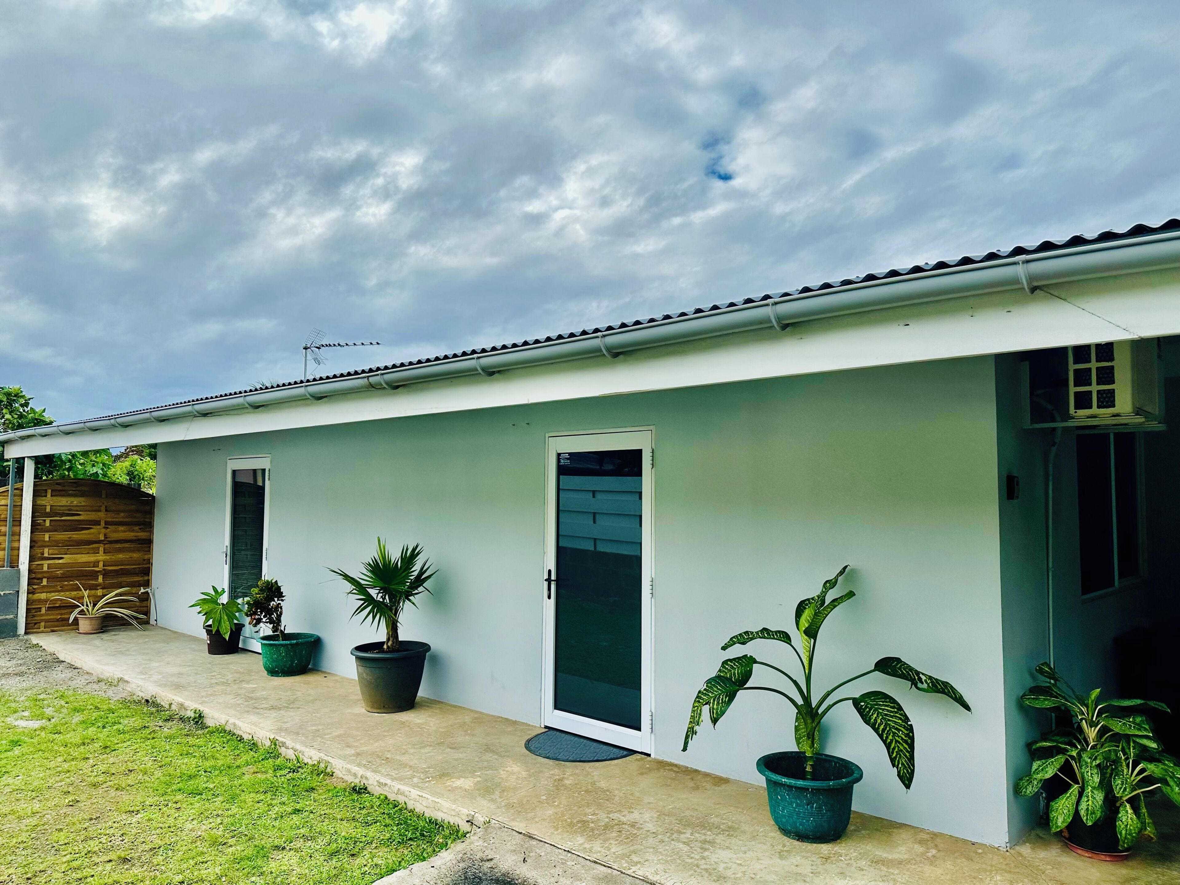 A single-story building with a flat roof, a front door, and a small porch area with potted plants.