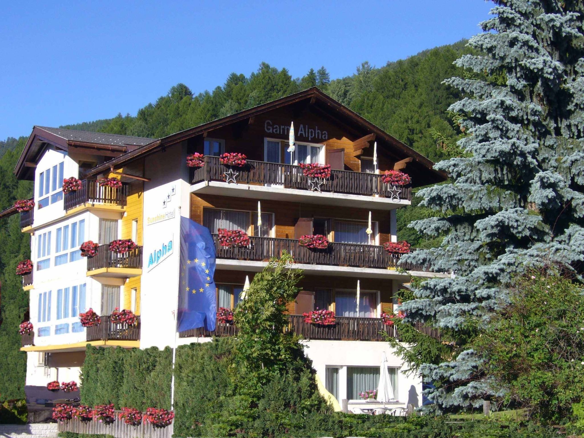 A multi-story hotel with balconies adorned with flowers, a visible flag, and a mountainous backdrop.
