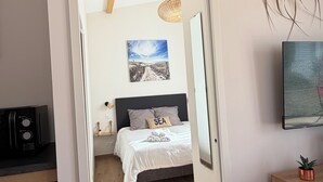 Ein Schlafzimmer mit einem Bett, einem Wandbild, einem kleinen Tisch mit einer Pflanze und Blick auf den Strand durch eine offene Tür.