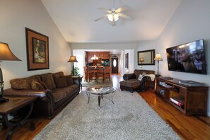 A living room with a sofa, coffee table, and TV. There is a dining area with chairs and a table. The room has wooden flooring and a ceiling fan. - Riverfront home near Dollywood and Pigeon Forge Parkway! (PIGEON FORGE)