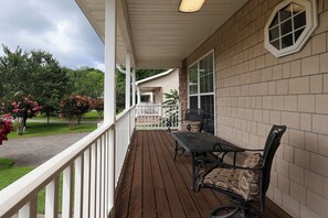 A wooden deck with a table and chairs, a white railing, and a view of a garden with trees and flowers. - Riverfront home near Dollywood and Pigeon Forge Parkway! (PIGEON FORGE)