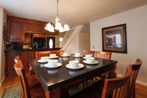 Comedor con mesa negra dispuesta para seis, sillas de madera, una araña y una cocina al fondo.